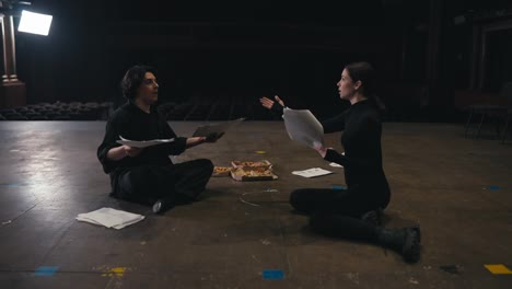 A-couple-of-confident-actors-a-brunette-girl-in-a-black-uniform-and-a-guy-with-a-kit-of-papers-and-a-script-play-their-roles-and-communicate-about-the-script-while-sitting-on-stage-in-an-empty-theater