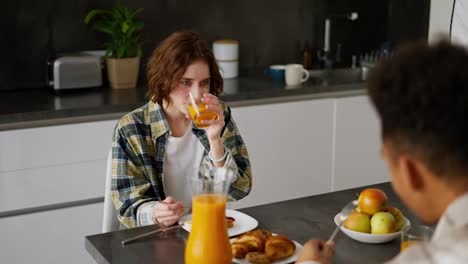 Over-the-shoulder-a-happy-young-adult-girl-with-brown-hair-and-a-bob-hairstyle-in-green-checkered-is-having-breakfast-with-her-boyfriend-with-Black-brunette-skin-color-during-a-shared-breakfast-in-the-morning-in-the-kitchen