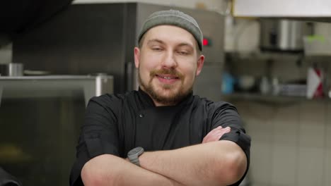 Retrato-De-Un-Chef-Feliz-Con-Barba-Y-Uniforme-Negro-Que-Cruza-Los-Brazos-Sobre-El-Pecho-Y-Posa-Para-La-Cámara-En-La-Cocina-De-Un-Restaurante.