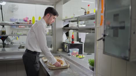 A-confident-guy-waiter-in-a-white-shirt-takes-the-order-and-puts-it-in-a-cardboard-pizza-box-tying-it-with-a-neat-bow.-Waiter-packing-a-dish-for-delivery