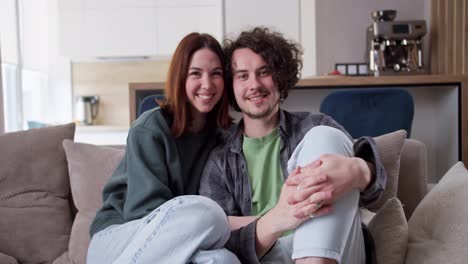 Portrait-of-a-happy-couple-brunette-girl-hugging-her-boyfriend-with-curly-hair-in-a-plaid-shirt-while-sitting-on-the-sofa-at-home-in-a-modern-studio-apartment