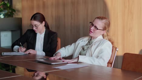 A-confident-and-happy-blonde-girl-with-glasses-and-a-white-business-suit-closes-her-laptop-and-leans-on-the-back-of-her-chair-during-success-and-victory-at-the-end-of-the-working-day-in-a-modern-office