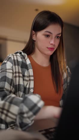 Vertical-video-of-a-confident-and-happy-brunette-girl-in-a-checkered-shirt-and-an-orange-T-shirt-working-on-her-laptop-in-a-modern-apartment-in-the-evening