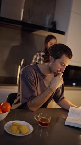 Vertical-video-scene-setting-of-a-brunette-man-in-a-gray-T-shirt-reading-a-book-after-dinner-while-his-brunette-girlfriend-in-a-checkered-shirt-plays-with-a-kitchen-knife-in-a-modern-apartment.-Scene-from-horror-films-a-brunette-girl-plays-with-a-knife-near-her-boyfriend