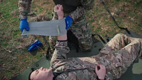 A-confident-male-military-man-in-a-camouflage-green-uniform-ties-a-fabric-bandage-to-the-unconscious-hand-of-a-male-soldier-in-a-combat-area-outside-the-city