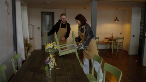 Happy-brunette-girl-waiter-in-a-yellow-apron-together-with-a-fellow-guy-putting-chairs-on-the-table-while-preparing-for-cleaning-and-ending-the-day-in-a-cafe