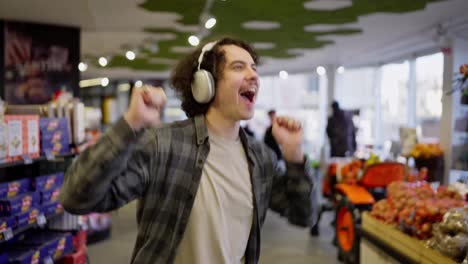 Cheerful-brunette-guy-in-a-checkered-shirt-wearing-white-headphones-listens-to-music-and-dances-in-a-grocery-store