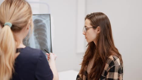 A-confident-young-brunette-girl-in-round-glasses-and-a-plaid-shirt-talks-to-a-blonde-girl-doctor-about-chest-fluorography-in-a-modern-clinic.-Over-the-shoulder-of-a-girl-Doctor-shows-the-results-of-fluorography-on-pictures-of-the-chest-of-a-young-brunette-girl