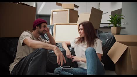 A-brunette-man-with-stubble-in-a-red-hat-and-a-beige-T-shirt-is-talking-with-his-brunette-girlfriend-in-a-white-T-shirt-sitting-on-the-floor-leaning-on-the-sofa-in-a-black-plastic-cover-and-among-a-huge-number-of-boxes-in-a-new-apartment-after-moving