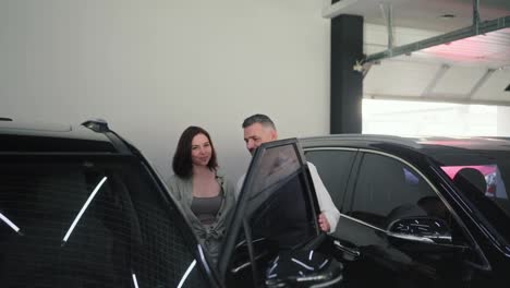 A-confident-middle-aged-man-with-a-gray-beard-and-wearing-a-white-shirt-is-communicating-with-his-brunette-wife-and-their-little-son-about-a-car-in-a-modern-car-dealership