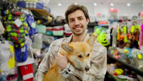 Portrait-of-a-happy-brunette-guy-in-a-plaid-shirt-who-holds-his-yellow-corgi-dog-in-his-arms-in-a-pet-store