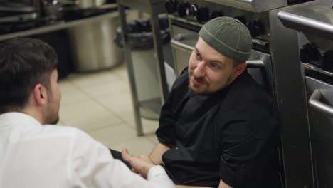 A-confident-professional-chef-in-a-black-uniform-with-a-beard-communicates-with-a-young-guy-waiter-who-supports-and-pats-the-chef-on-the-shoulder-while-sitting-on-the-floor-in-the-kitchen-in-a-restaurant