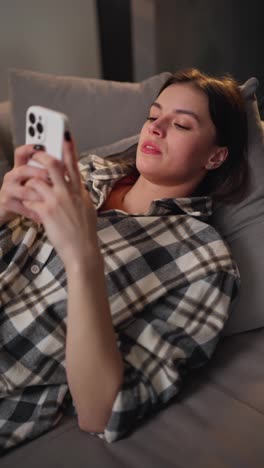 Vertical-video-of-an-enraged-and-irritated-brunette-girl-in-a-checkered-shirt-lies-on-a-gray-sofa-and-communicates-using-her-new-smartphone-in-a-modern-apartment