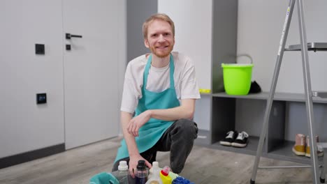 Portrait-of-a-confident-blond-man-in-a-white-T-shirt-as-a-cleaner-in-a-blue-apron-who-sits-on-the-floor-and-poses-near-cleaning-supplies-collected-in-a-gray-plastic-basin-and-while-cleaning-on-call-in-a-modern-apartment