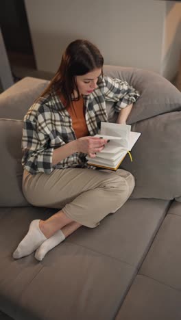 Vertical-video-from-above-of-a-confident-and-calm-brunette-girl-in-a-checkered-shirt-and-an-orange-T-shirt-sitting-on-a-gray-sofa-and-reading-a-book-at-home-in-a-modern-apartment