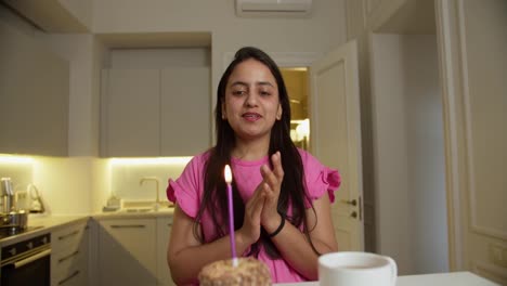 Happy-brunette-girl-in-a-pink-dress-claps-her-hands-and-blows-out-a-pink-candle-on-her-small-cake-while-sitting-at-a-table-in-a-modern-apartment