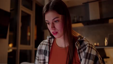 Close-up-of-a-confident-brunette-girl-in-a-checkered-shirt-and-an-orange-T-shirt-watching-funny-videos-while-sitting-on-the-sofa-in-a-modern-apartment-in-the-evening