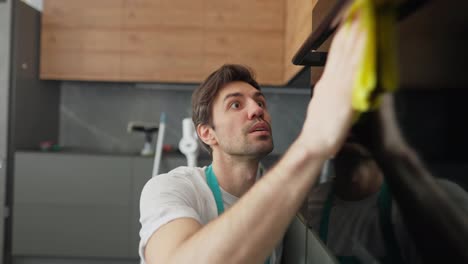 A-confident-brunette-guy-in-a-white-T-shirt-and-a-blue-apron-is-a-cleaner-washing-a-glossy-surface-with-a-yellow-rag-and-detergent-in-the-kitchen-in-a-modern-apartment