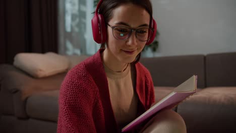 Close-up-of-a-confident-brunette-girl-with-glasses-in-a-red-sweater-and-wireless-headphones-studying-foreign-languages-online-and-taking-notes-in-her-notebook-in-a-modern-apartment