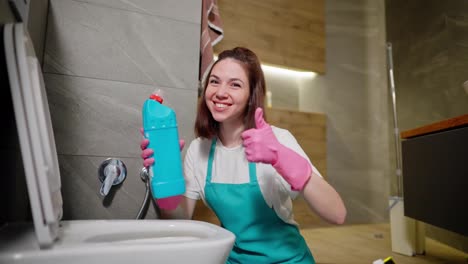 Retrato-De-Una-Alegre-Y-Segura-Señora-De-La-Limpieza-Morena-Con-Una-Camiseta-Blanca-Y-Un-Delantal-Azul-Señala-El-Detergente-En-Su-Mano,-Forja-Su-Cabeza-Y-Le-Da-Un-Me-Gusta-En-Un-Guante-De-Goma-Rosa-En-El-Baño-De-Un-Apartamento-Moderno-Mientras-Limpia.