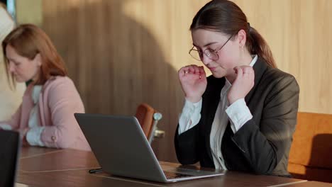 Close-up-of-a-tired-brunette-girl-in-business-clothes-loses-her-temples-and-adjusts-her-glasses-while-working-in-front-of-a-laptop-in-a-modern-sunny-office