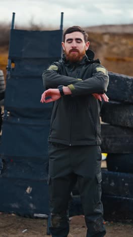 Vertical-video-of-a-confident-brunette-man-with-a-beard-in-a-dark-green-army-jacket-and-pants-standing-near-tires-in-the-steppe-and-posing.-Portrait-of-a-confident-military-man-standing-near-tires-during-a-training-exercise