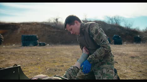 A-confident-young-man-in-army-camouflage-and-blue-medical-gloves-puts-a-thick-fabric-bandage-on-the-hand-of-his-teammate-during-combat-operations-in-the-steppe-territory