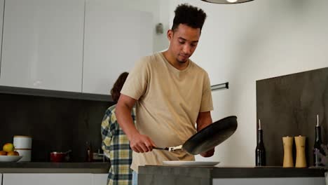 A-young-Black-skinned-brunette-man-in-a-beige-T-shirt-puts-scrambled-eggs-on-a-plate-while-they-are-preparing-breakfast-together-with-his-girlfriend-a-young-adult-brunette-who-washes-the-dishes-they-are-preparing-for-breakfast-and-in-the-morning-in-a-modern-apartment