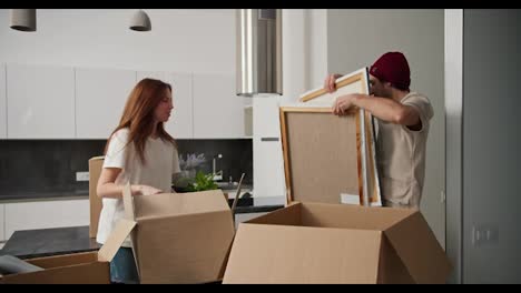 Happy-man-with-stubble-in-a-red-cap-gives-his-brunette-girlfriend-in-a-white-T-shirt-things-from-Big-Boxes-during-his-move-in-a-modern-apartment