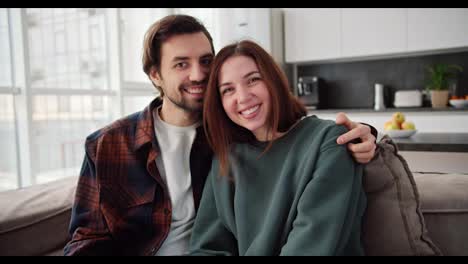 Portrait-of-a-happy-couple,-a-brunette-girl-in-a-green-sweater-together-with-her-brunette-boyfriend-with-stubble-in-a-checkered-shirt-posing-while-sitting-on-the-sofa-in-a-modern-apartment