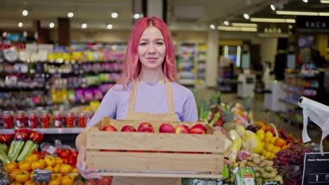 Portrait-of-a-girl-with-pink-hair-in-an-apron-posing-with-a-box-of-apples-while-working-in-a-supermarket