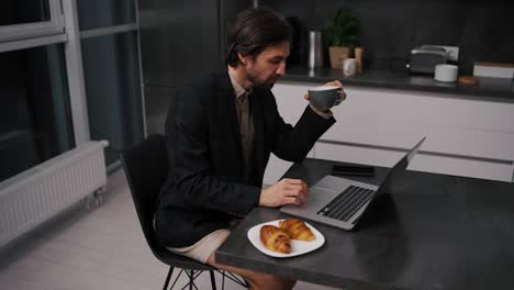 A-confident-brunette-man-with-stubble-in-a-black-jacket-works-remotely-and-communicates-using-a-laptop-while-sitting-on-a-chair-in-beige-shorts-in-the-evening-in-a-modern-apartment