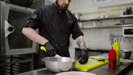 Close-up-of-a-confident-male-chef-in-a-black-uniform-with-a-bandana-and-protective-gloves-cutting-a-salad-and-putting-it-into-a-bowl-while-preparing-a-dish-in-a-modern-kitchen-in-a-restaurant