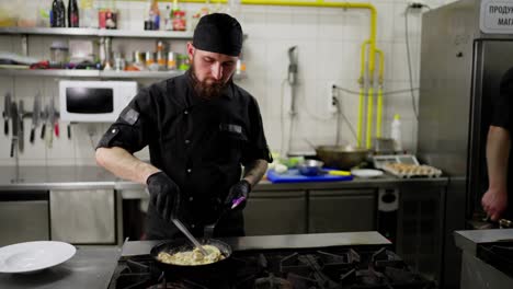 A-professional-male-chef-with-a-beard-in-a-black-uniform-and-with-a-bandana-stirs-food-in-a-frying-pan-while-preparing-a-dish-in-a-restaurant-kitchen