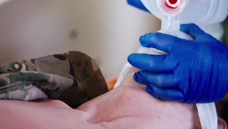 Close-up-a-confident-male-military-medic-in-blue-rubber-gloves-pumps-air-using-a-matte-white-bottle-and-performs-artificial-respiration-on-an-unconscious-wounded-soldier-in-an-ambulance