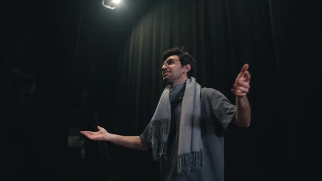 A-confident-brunette-man-in-a-gray-T-shirt-and-scarf-the-stage-director-goes-on-stage-and-greets-everyone-in-the-theater-with-black-curtains