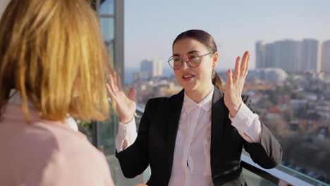 Over-the-shoulder-a-confident-brunette-girl-in-round-glasses-and-a-black-business-uniform-communicates-with-her-colleague-a-blonde-girl-with-a-bob-hairstyle-while-relaxing-on-the-terrace-while-working-overlooking-the-city-in-sunny-weather-in-the-evening