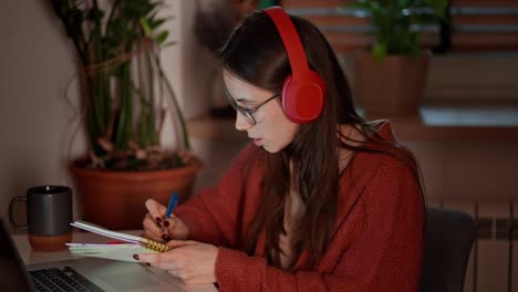 Confident-brunette-girl-in-wireless-headphones-and-glasses-takes-notes-in-her-notes-while-learning-foreign-languages-using-online-lessons-on-a-laptop-while-sitting-in-a-modern-kitchen-in-an-apartment