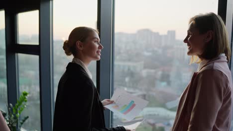 Confident-young-blonde-girl-in-a-business-uniform-shows-her-papers-and-research-on-the-work-of-her-colleague-while-communicating-near-panoramic-windows-overlooking-the-city-in-a-modern-office