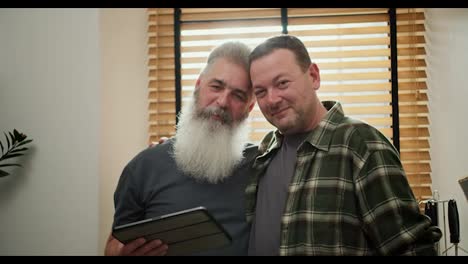 Retrato-De-Una-Feliz-Pareja-De-Dos-Hombres-Homosexuales-Lgbt,-Un-Hombre-De-Mediana-Edad-Con-Cabello-Gris-Y-Barba-Incipiente-Con-Una-Camisa-A-Cuadros-Verde-Junto-Con-Su-Novio-Mayor-Con-Cabello-Gris-Y-Una-Exuberante-Barba-Gris-Con-Una-Camiseta-Gris-Que-Sostiene-Una-Tableta-Electrónica-En-La-Cocina-En-Un-Apartamento-Moderno