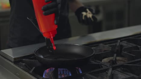 Close-up-of-a-male-professional-chef-in-a-black-uniform-and-gloves-puts-oil-on-a-frying-pan-and-adds-mushrooms-there-while-preparing-a-dish-on-a-gas-burner-in-a-restaurant-kitchen