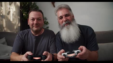 A-happy-brunette-man-with-stubble-in-a-gray-T-shirt-plays-video-games-using-a-joystick-with-his-boyfriend.-An-elderly-man-with-gray-hair-and-a-lush-beard-he-wins-and-rejoices.-Sitting-on-the-sofa-in-a-modern-apartment-in-the-cozy-evening