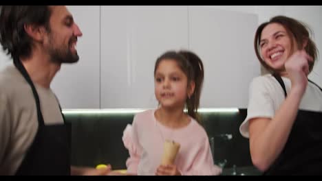 Close-up-of-a-happy-brunette-girl-in-pink-clothes-jumping-and-having-fun-with-her-parents-who-kiss-her-on-the-cheek-while-preparing-for-breakfast-in-the-morning-in-a-modern-apartment-in-the-kitchen