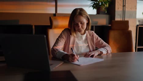 Portrait-of-a-confident-middle-aged-blonde-girl-in-a-pink-jacket-and-modern-uniform-who-sits-at-a-wooden-table-in-a-sunny-office-and-smiles