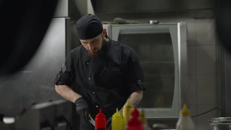 Side-view-of-a-confident-male-chef-in-a-black-uniform-wearing-black-rubber-gloves-preparing-a-salad-and-cutting-vegetables-in-the-kitchen-in-a-restaurant