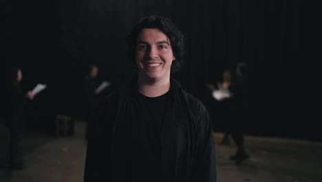 Portrait-of-a-young-confident-brunette-man-in-a-black-suit-the-actor-smiles-and-poses-near-his-colleagues-who-are-preparing-for-a-rehearsal-and-performance-on-stage-in-the-theater