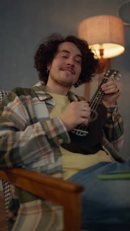 Vertical-close-up-video-of-a-happy-brunette-guy-with-curly-hair-and-a-mustache-in-a-checkered-shirt-playing-a-black-ukulele-near-a-lamp-during-an-atmospheric-evening