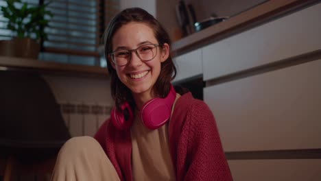 Retrato-De-Una-Joven-Morena-Feliz-Con-Gafas,-Auriculares-Inalámbricos-Rojos-Y-Un-Suéter-Que-Está-Sentada-Y-Posando-En-El-Suelo-De-La-Cocina-En-Un-Apartamento-Moderno-Por-La-Noche.