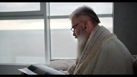 An-elderly-man-with-gray-hair-and-a-lush-beard-is-wrapped-in-a-white-blanket-looking-at-a-magazine-while-sitting-on-a-brown-sofa-in-a-modern-apartment-overlooking-the-sea