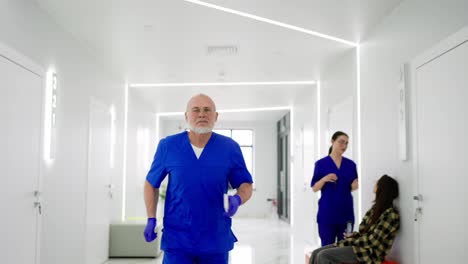 A-hurrying-older-man-doctor-in-a-blue-uniform-runs-along-the-bright-corridor-of-the-clinic-holding-a-phone-in-his-hands.-An-elderly-man-doctor-with-a-beard-runs-along-the-corridor-of-a-modern-on-call-clinic